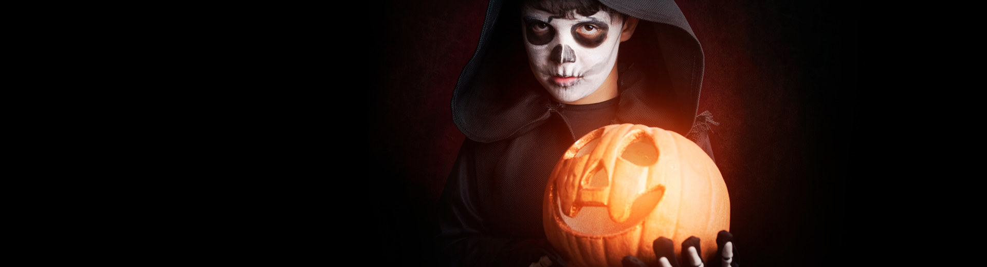 Person i sort kappe holder en utskåret, opplyst gresskarlykt med smilende ansikt.