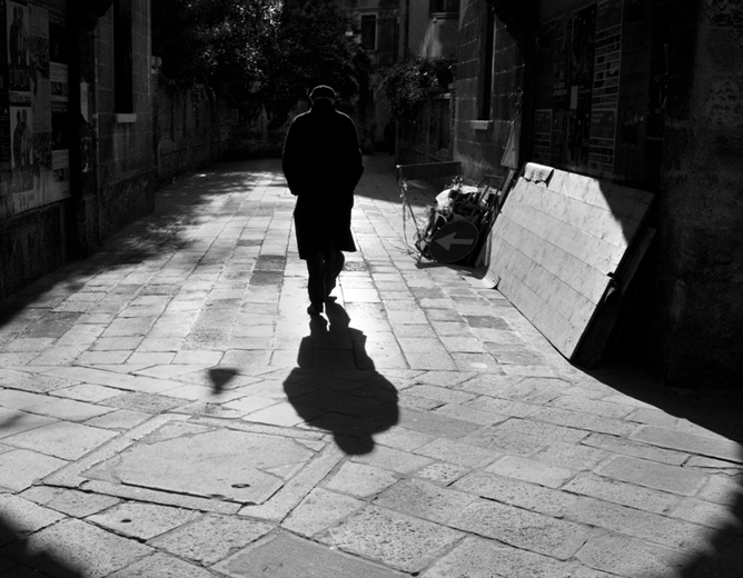 En person som går alene nedover en smal, solfylt brosteinsgate med lange skygger rundt seg
