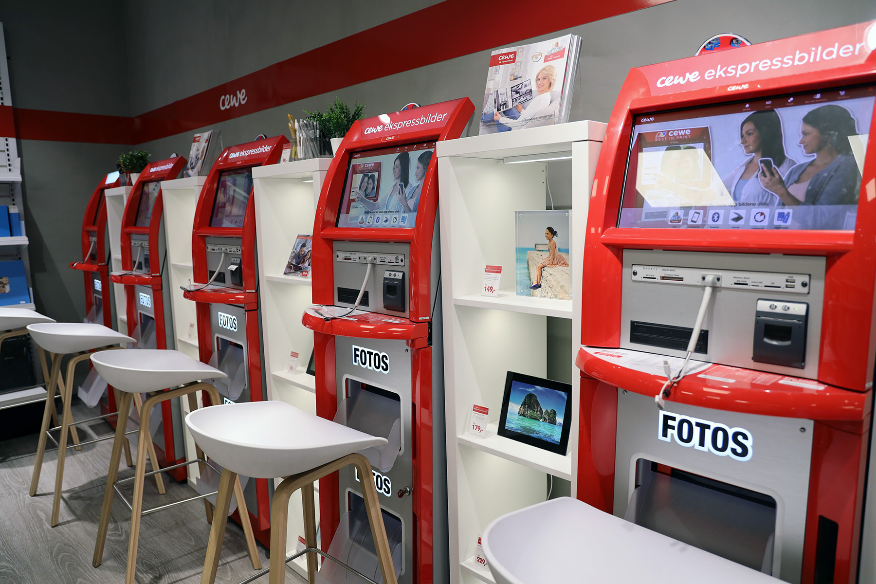 Røde CEWE fotokiosker med skjermer og skrivere plassert på en rad, omgitt av hvite hyller med innrammede bilder og dekor. Røde CEWE fotokiosker med skjermer og skrivere plassert på en rad, omgitt av hvite hyller med innrammede bilder og dekor.