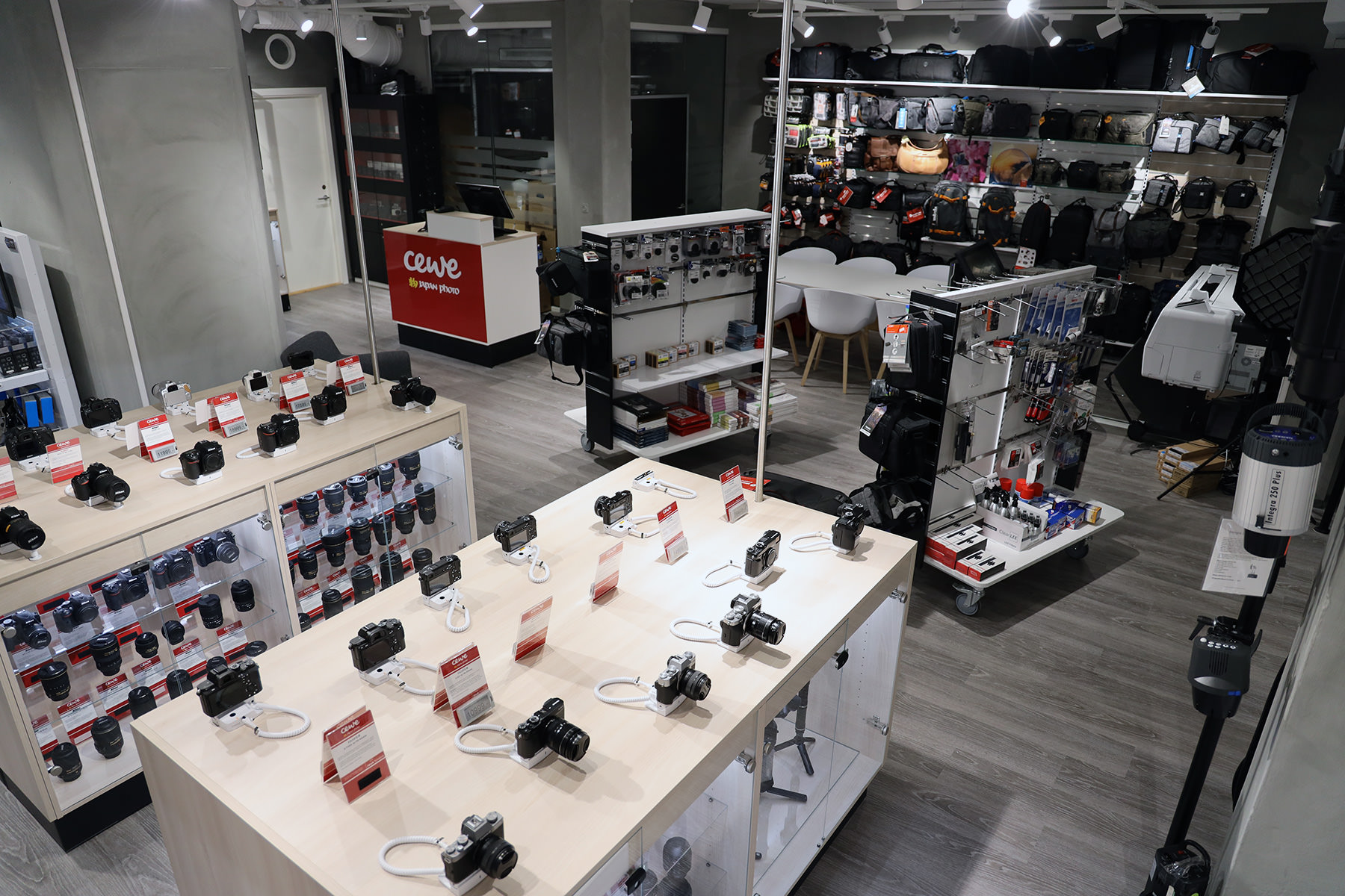 Butikkinteriør med kameraer og objektiver utstilt på lyse bord og hyller, vegger fylt med kameravesker og tilbehør, CEWE-logo ved kassen Butikkinteriør med kameraer og objektiver utstilt på lyse bord og hyller, vegger fylt med kameravesker og tilbehør, CEWE-logo ved kassen