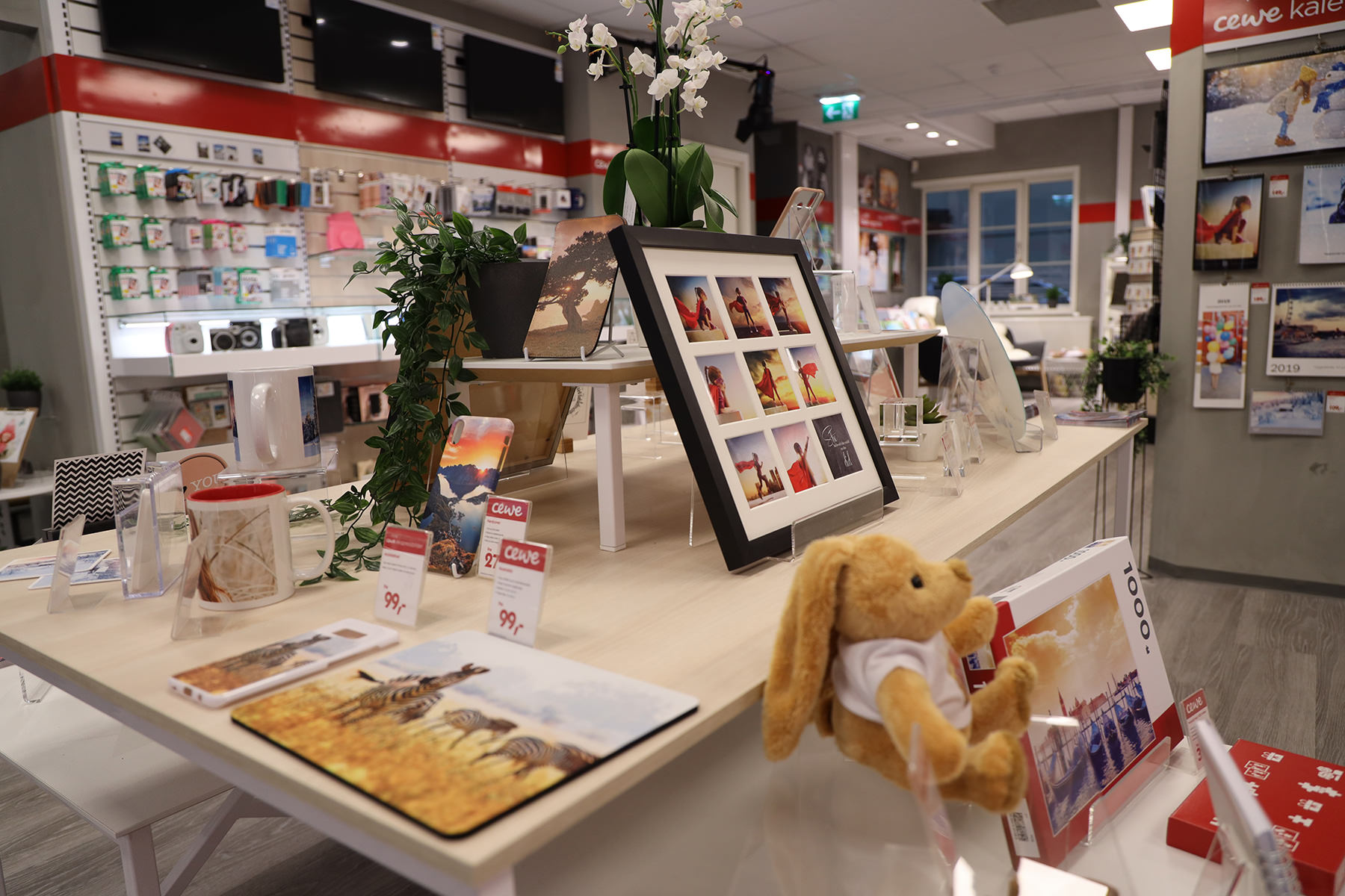 Utstilling med fotoprodukter, inkludert rammer, kopper, fotobøker og kosedyr, omgitt av planter og lyse interiørdetaljer Utstilling med fotoprodukter, inkludert rammer, kopper, fotobøker og kosedyr, omgitt av planter og lyse interiørdetaljer