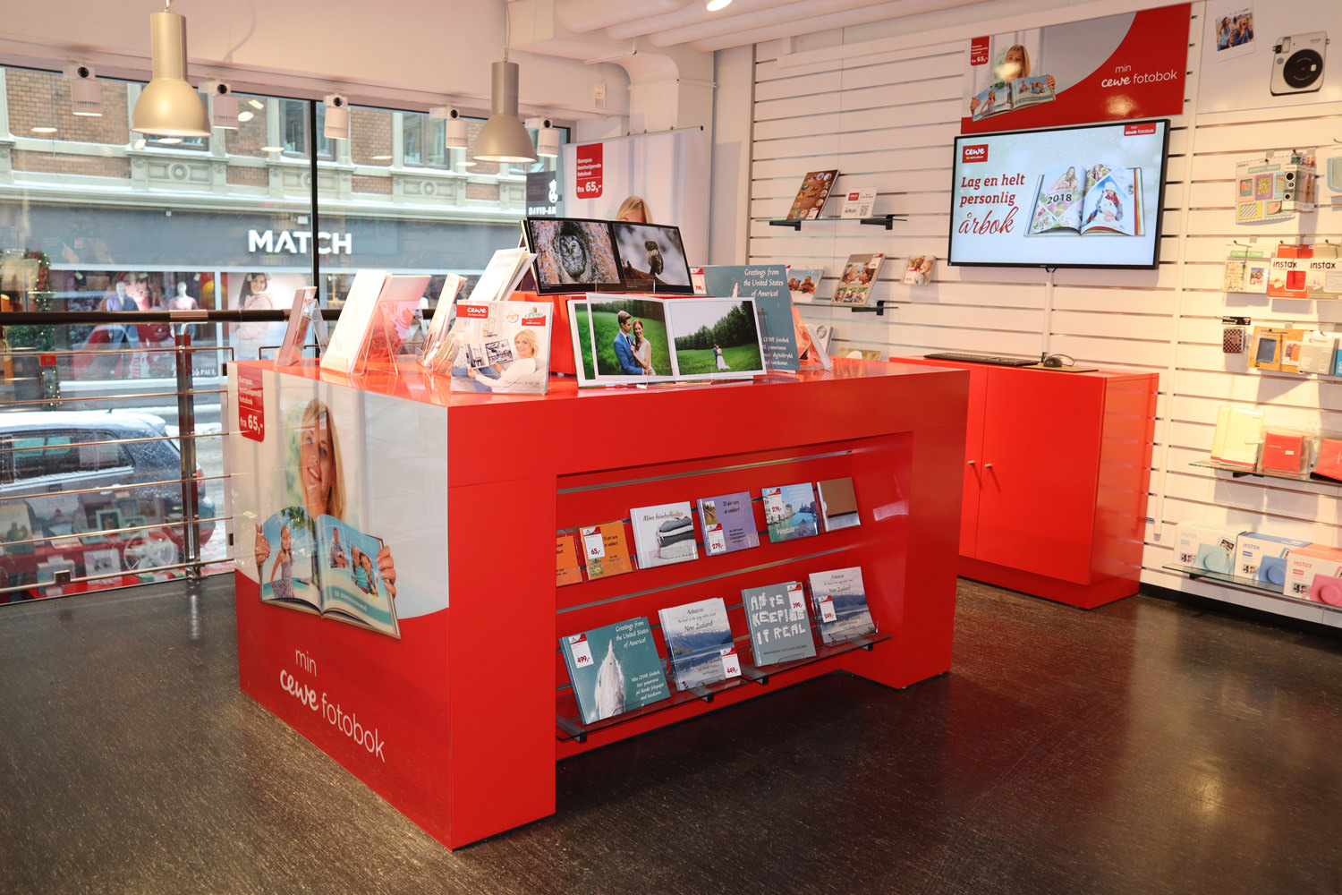 Rødt fotobok-display med CEWE-produkter på utstillingsbord og hyller i en lys butikk.