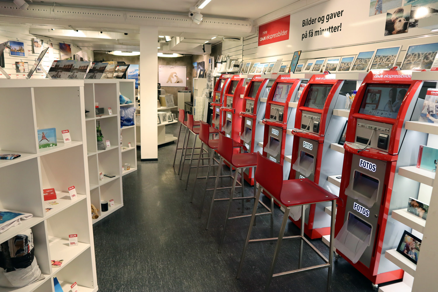 Røde selvbetjeningsautomater for fotoutskrifter med tilhørende stoler, omgitt av hyller med fotogaver og bilder