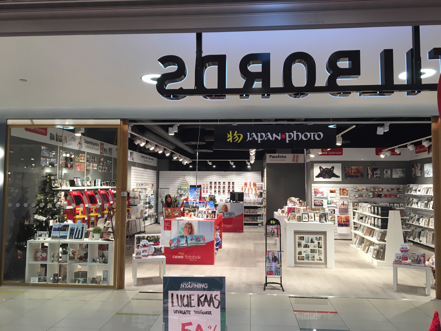Butikkinngang med skiltet "Japan Photo" i svart og rødt, dekorert med lys. Interiøret viser hyller med fotoprodukter, rammer og fotobøker, samt en tydelig rød dekorasjon med CEWE-logo.