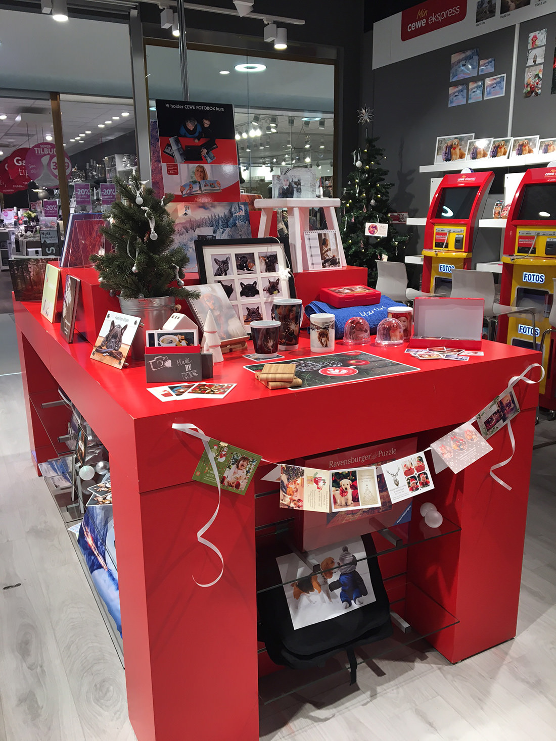 Rødt bord med ulike personlige fotoprodukter som kopper, album og kalendere, dekorert med juletema og små julekort.