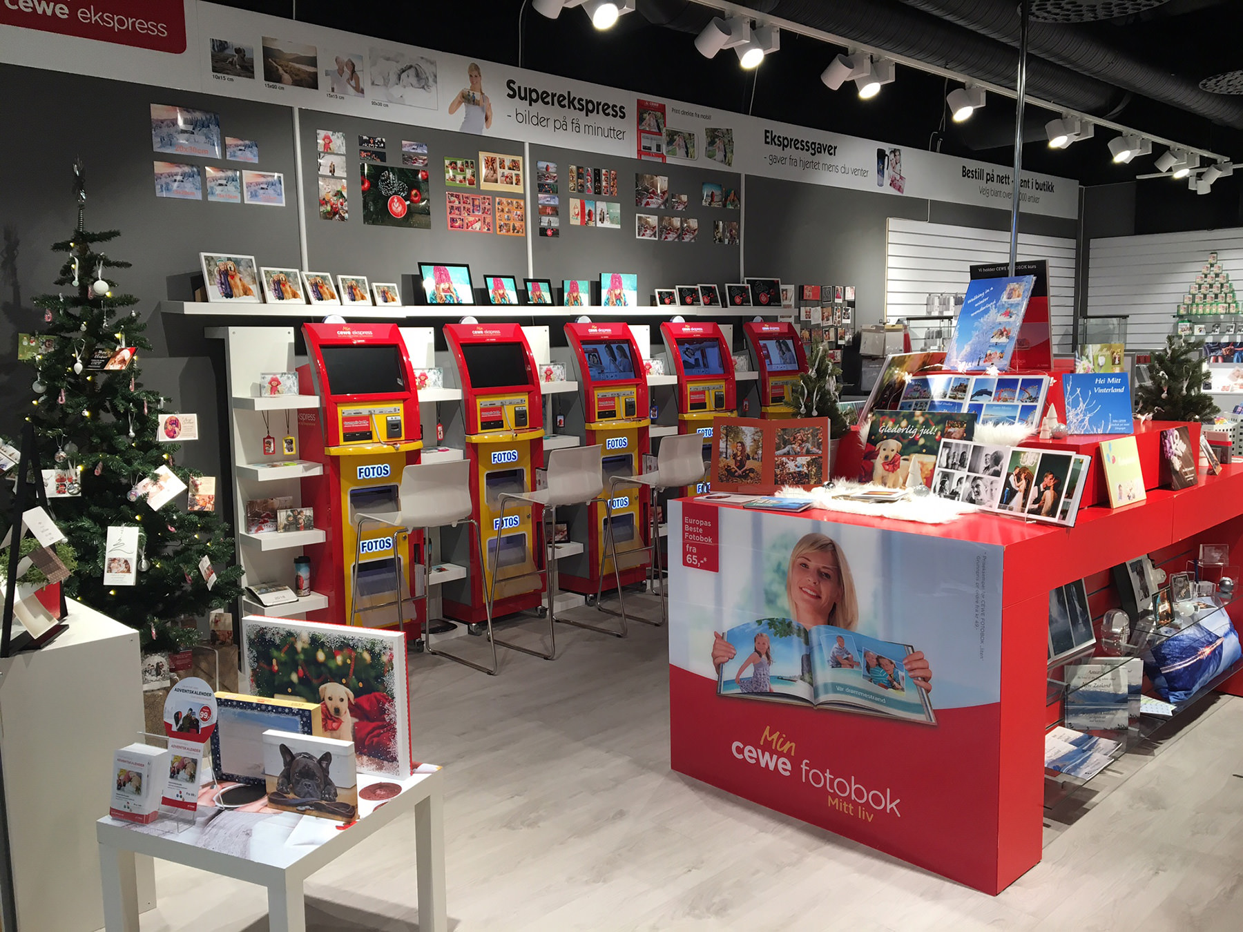 En CEWE-butikk med røde fotokiosker, juledekorasjoner og utstilte fotoprodukter som kort, bøker og kalendere