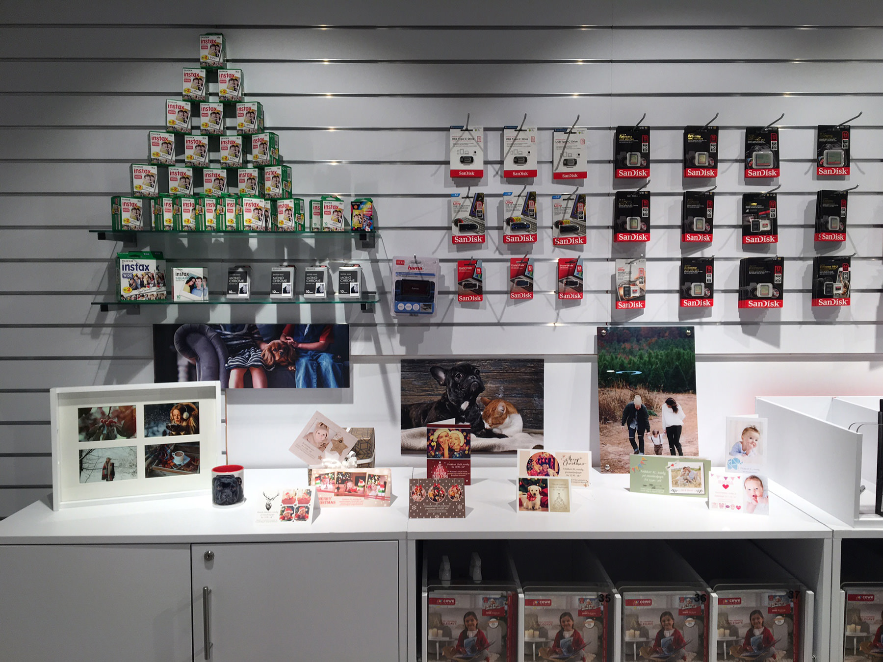 Utstilling med Instax-filmpakker formet som et juletre, SanDisk-minnekort på veggen, fotografier i rammer og fotogaver på en hylle.