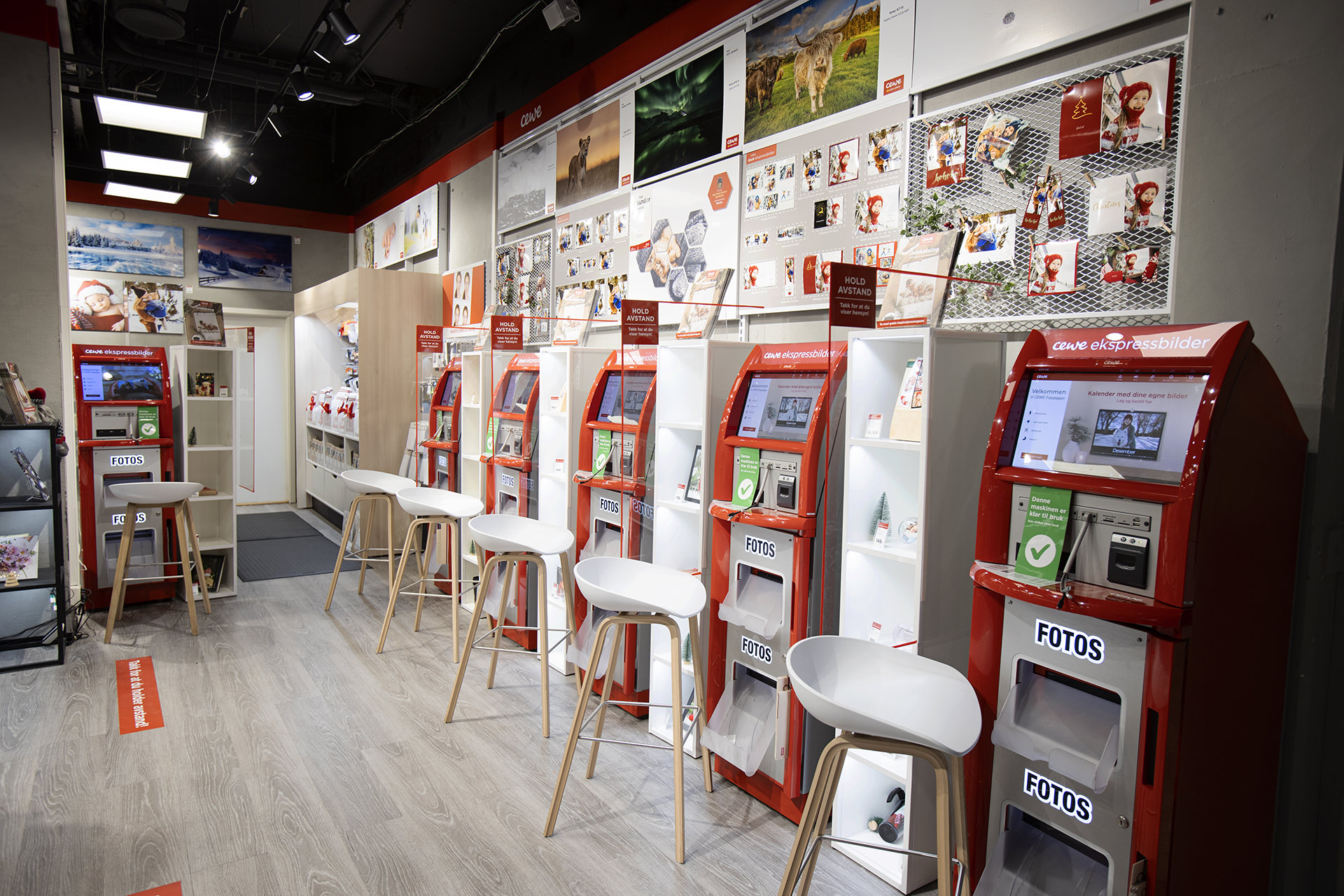 En lys og moderne butikk med CEWE fotokiosker på rekke, omgitt av høye hvite hyller med fotoprodukter og dekorasjoner. En lys og moderne butikk med CEWE fotokiosker på rekke, omgitt av høye hvite hyller med fotoprodukter og dekorasjoner.