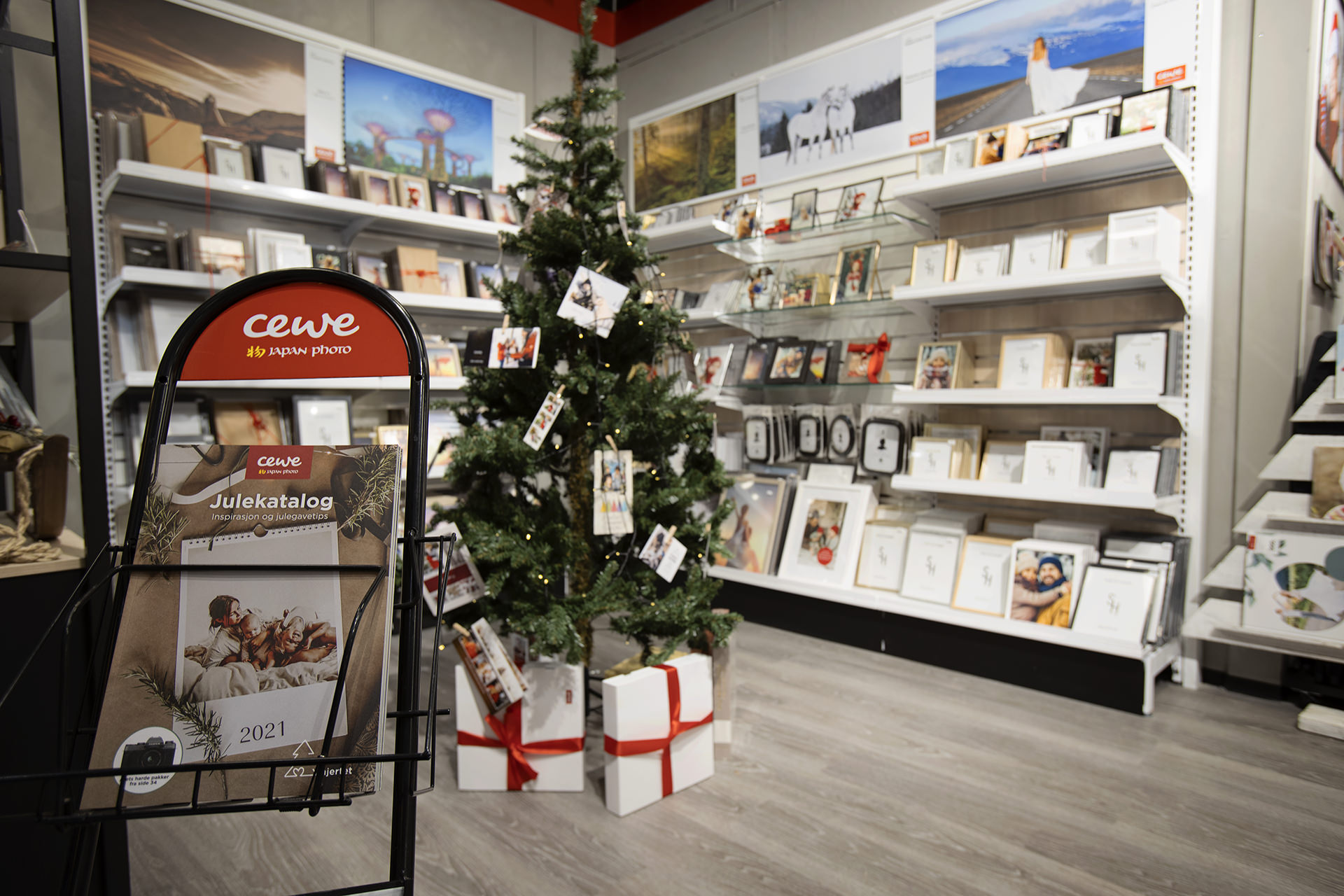 CEWE Julekatalog plassert i en svart brosjyreholder foran et pyntet juletre med gaver, omgitt av bilderammer og dekorasjoner CEWE Julekatalog plassert i en svart brosjyreholder foran et pyntet juletre med gaver, omgitt av bilderammer og dekorasjoner