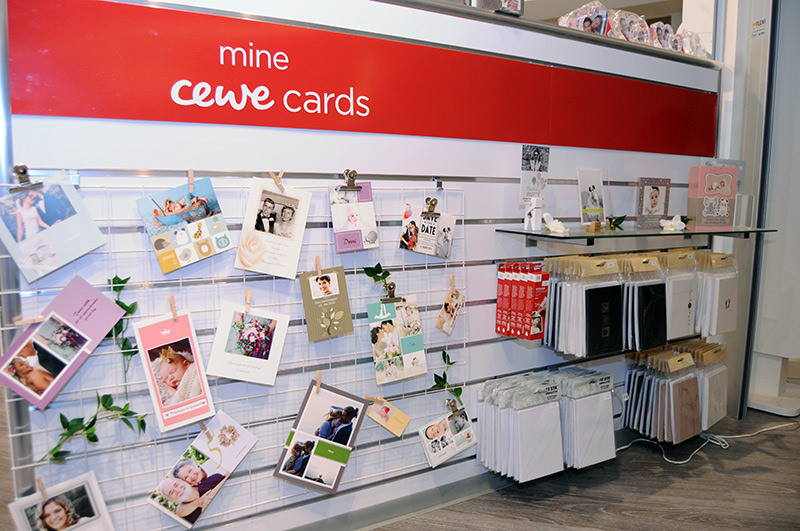 Stativ med et utvalg av personlige CEWE kort, festet på et gitter og hyller, dekorert med grønne blader. Stativ med et utvalg av personlige CEWE kort, festet på et gitter og hyller, dekorert med grønne blader.