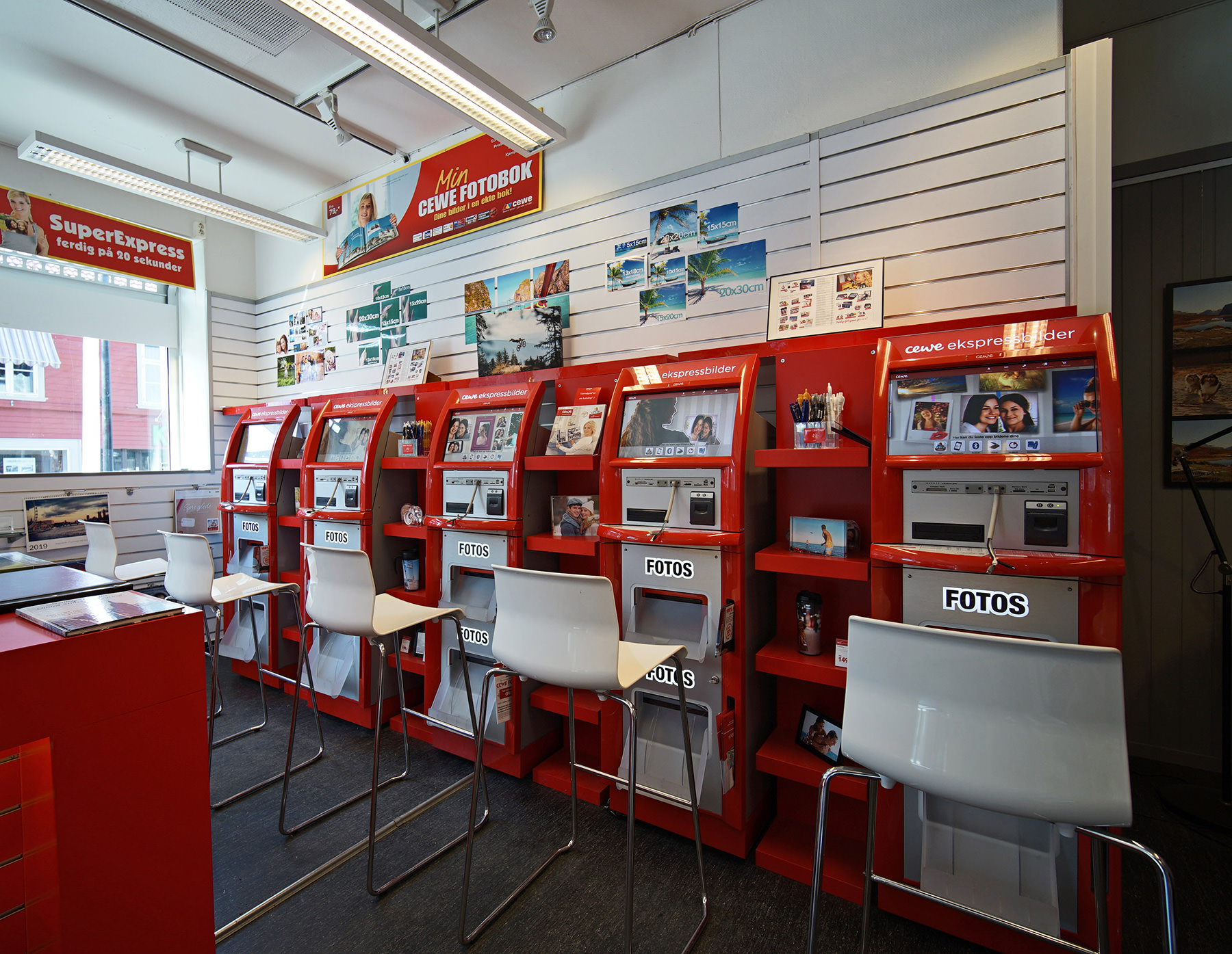 Selvbetjeningsområde med røde CEWE fotostasjoner og hvite stoler i en lys fotobutikk