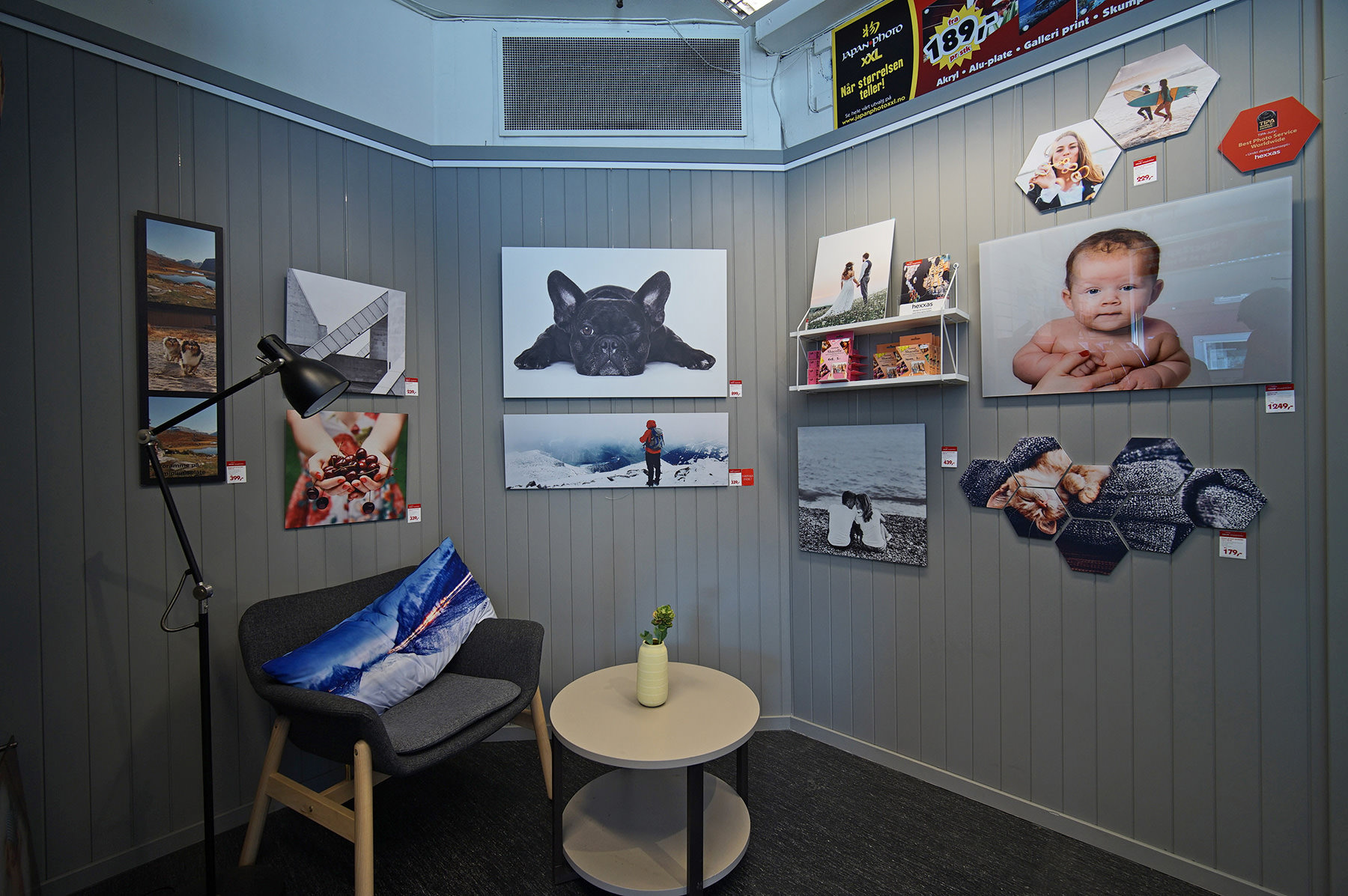 Utstillingsområde med lysegrå vegger dekorert med ulike fotoprodukter, inkludert lerretsbilder, sekskantede veggpaneler og en dekorativ pute på en grå stol.