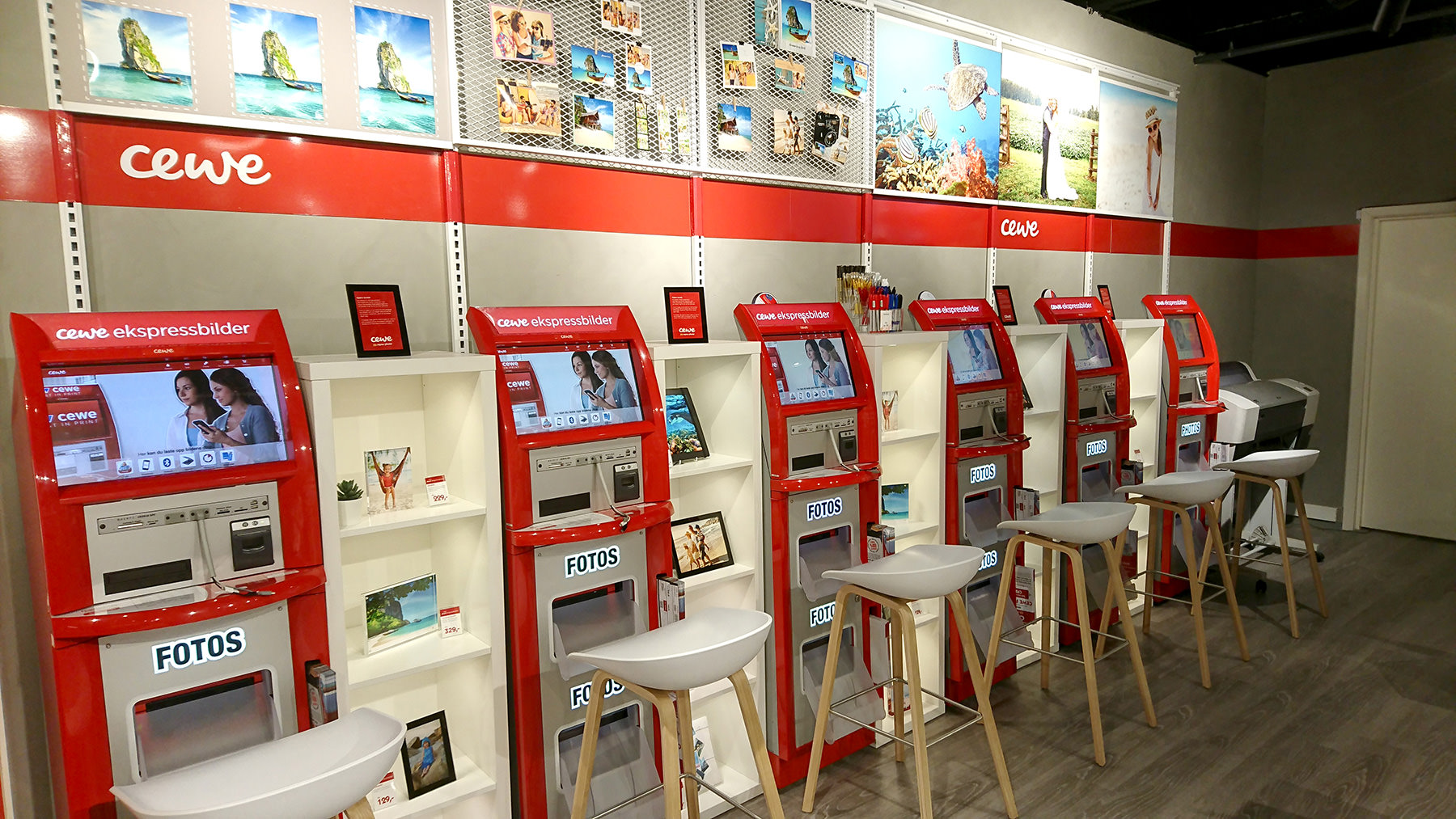 Røde CEWE fotokiosker på rad med skjermer, hyller og stoler i en moderne butikkinnredning Røde CEWE fotokiosker på rad med skjermer, hyller og stoler i en moderne butikkinnredning