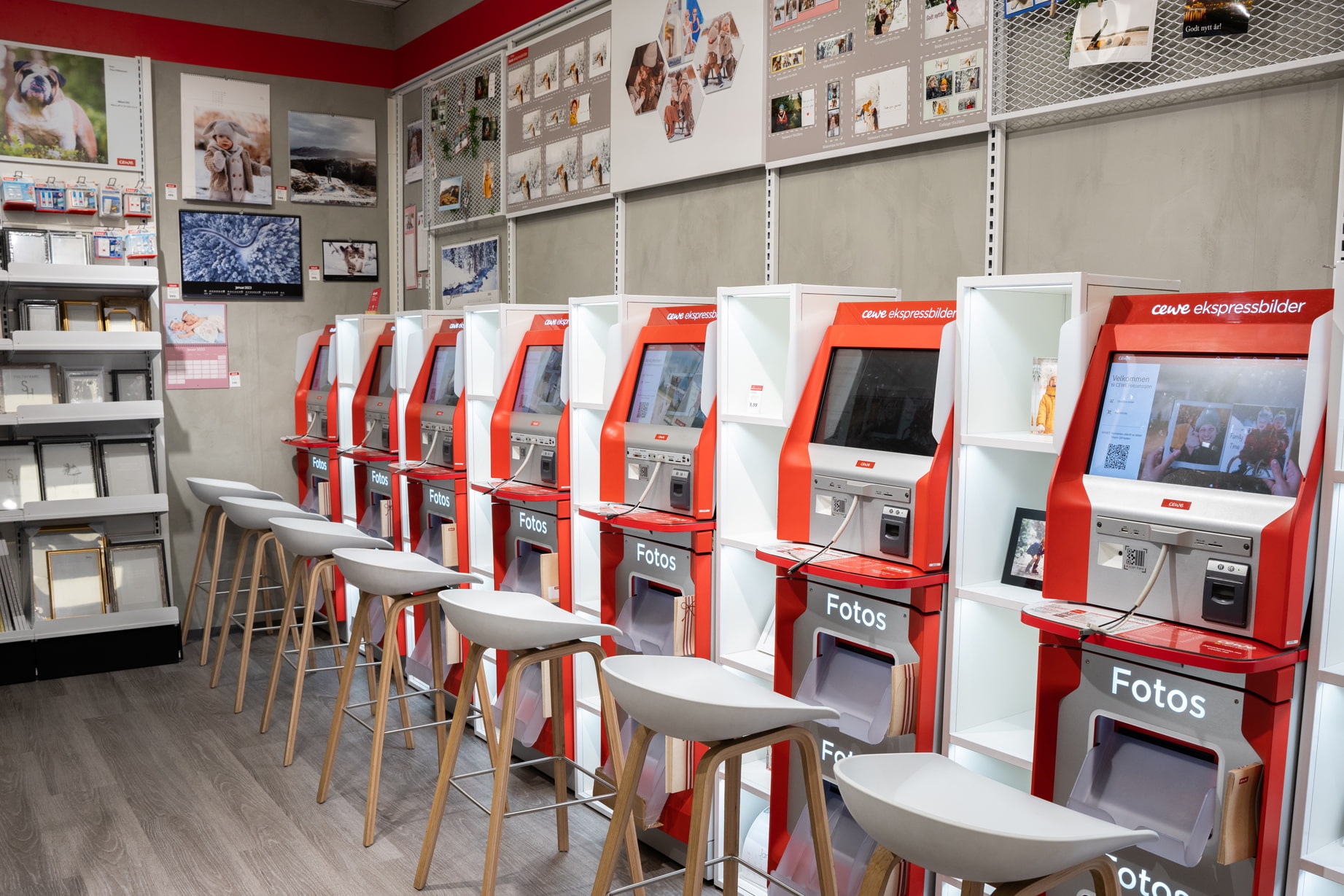 Røde CEWE fotostasjoner på rad med hvite barstoler foran, vegg dekorert med fotoprodukter