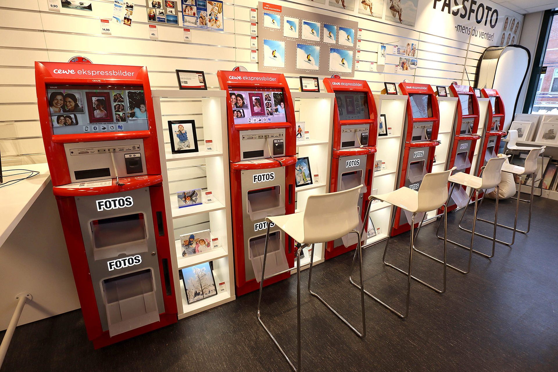Røde fotostasjoner med CEWE-logo foran en vegg med utstilte rammer og fotoprodukter Røde fotostasjoner med CEWE-logo foran en vegg med utstilte rammer og fotoprodukter
