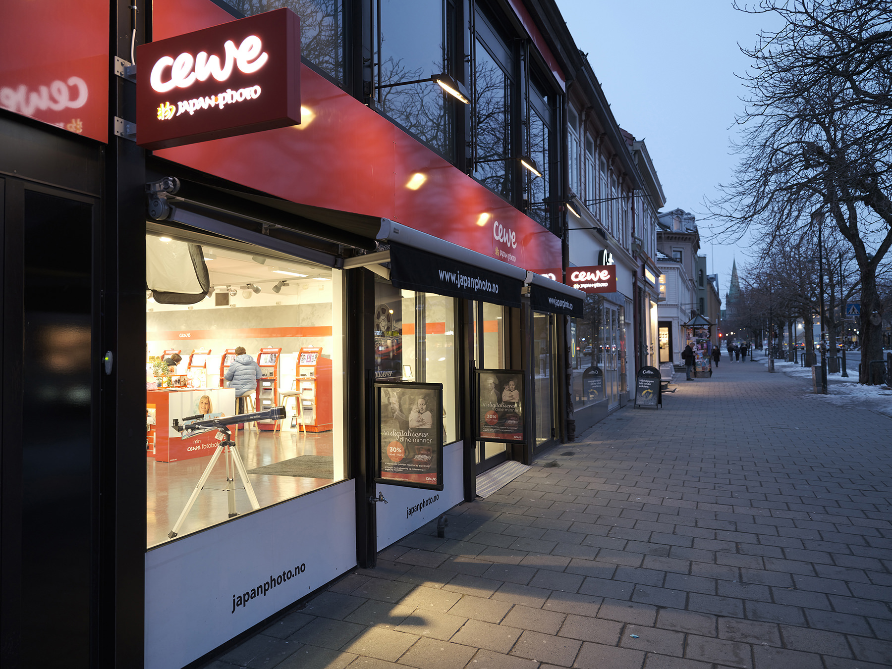 Butikkfasade til CEWE Japan Photo med store vinduer og belyst interiør, sett fra gaten i skumringen. Butikkfasade til CEWE Japan Photo med store vinduer og belyst interiør, sett fra gaten i skumringen.