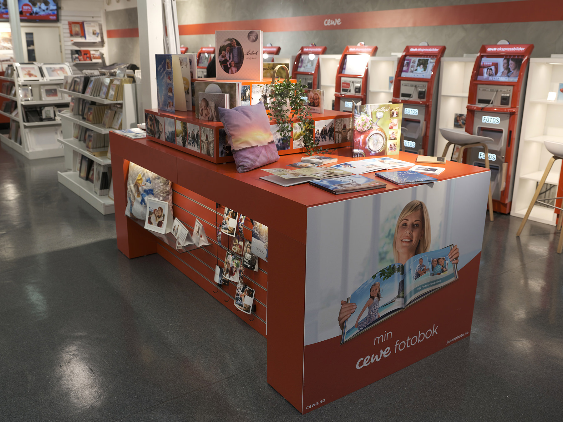 CEWE-butikk med utstillingsbord som viser fotobøker, kort og fotoprodukter, omgitt av fotokiosker og hyller med fotoutskrifter og rammer. CEWE-butikk med utstillingsbord som viser fotobøker, kort og fotoprodukter, omgitt av fotokiosker og hyller med fotoutskrifter og rammer.