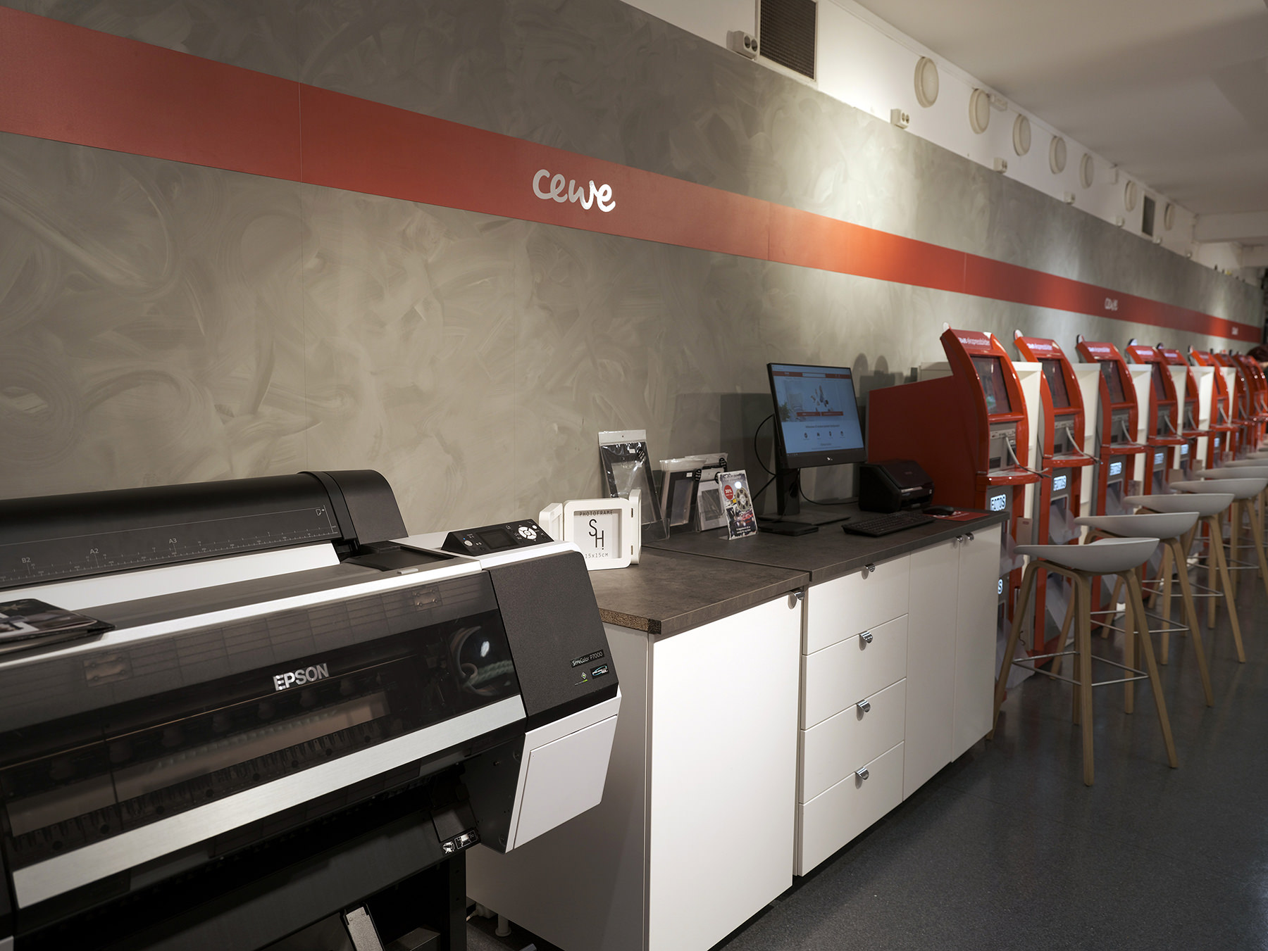 En rekke røde CEWE-terminaler med skjermer og krakker, Epson-skriver og hvite skap på en moderne grå bakgrunn En rekke røde CEWE-terminaler med skjermer og krakker, Epson-skriver og hvite skap på en moderne grå bakgrunn