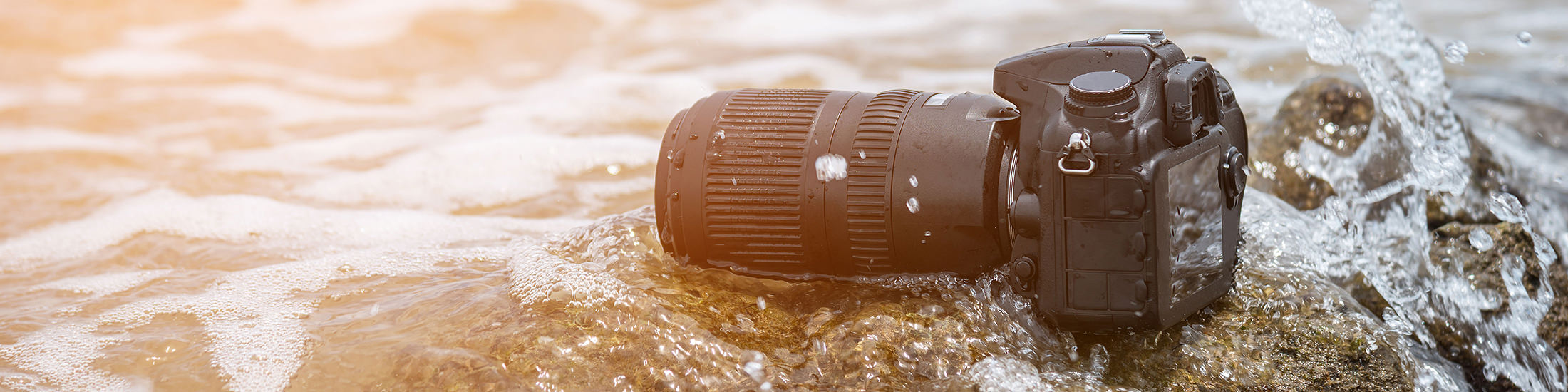 Kamera med stort objektiv ligger delvis i vann på en stein ved strandkant