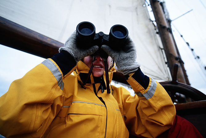 Person iført gul regnjakke og grå votter ser gjennom kikkert på en seilbåt   Person iført gul regnjakke og grå votter ser gjennom kikkert på en seilbåt