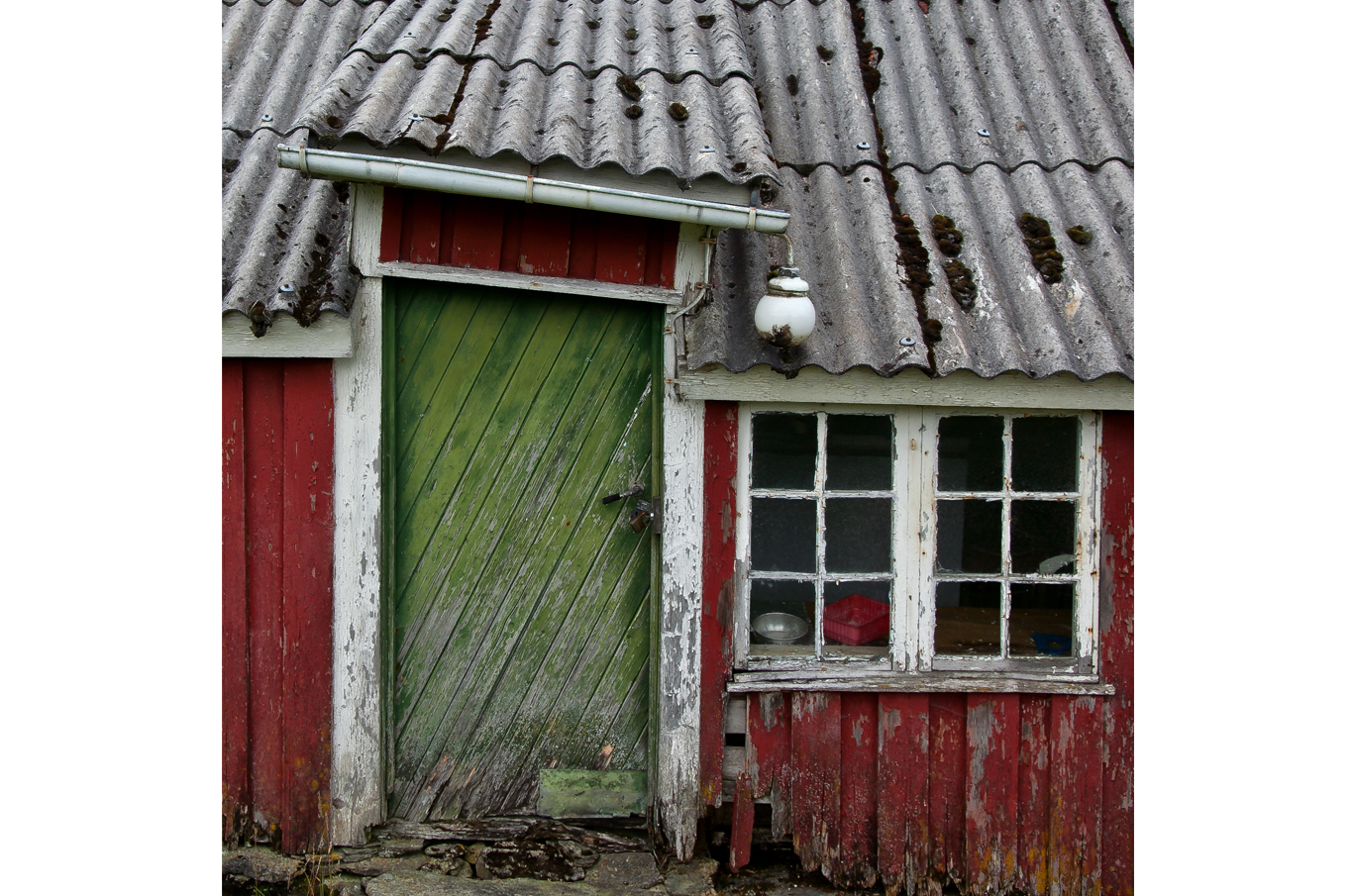 Slitt, grønn trefarget dør og hvitt vindu på en gammel, rød trehytte med bølgete tak av grå asbest Slitt, grønn trefarget dør og hvitt vindu på en gammel, rød trehytte med bølgete tak av grå asbest