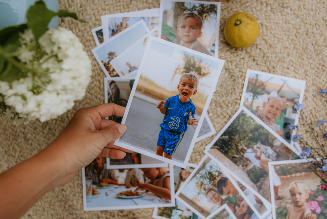 En hånd som holder et foto av et barn som løper utendørs, omgitt av flere spredte fotografier på en lys teppebakgrunn