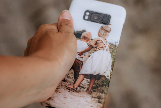 Hånd holder en mobiltelefon med et personlig deksel som viser to barn som går hånd i hånd i naturlige omgivelser
