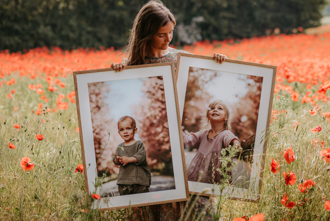 En jente i en blomstrende valmuemark holder to innrammede portrettfotografier med varme, myke fargetoner