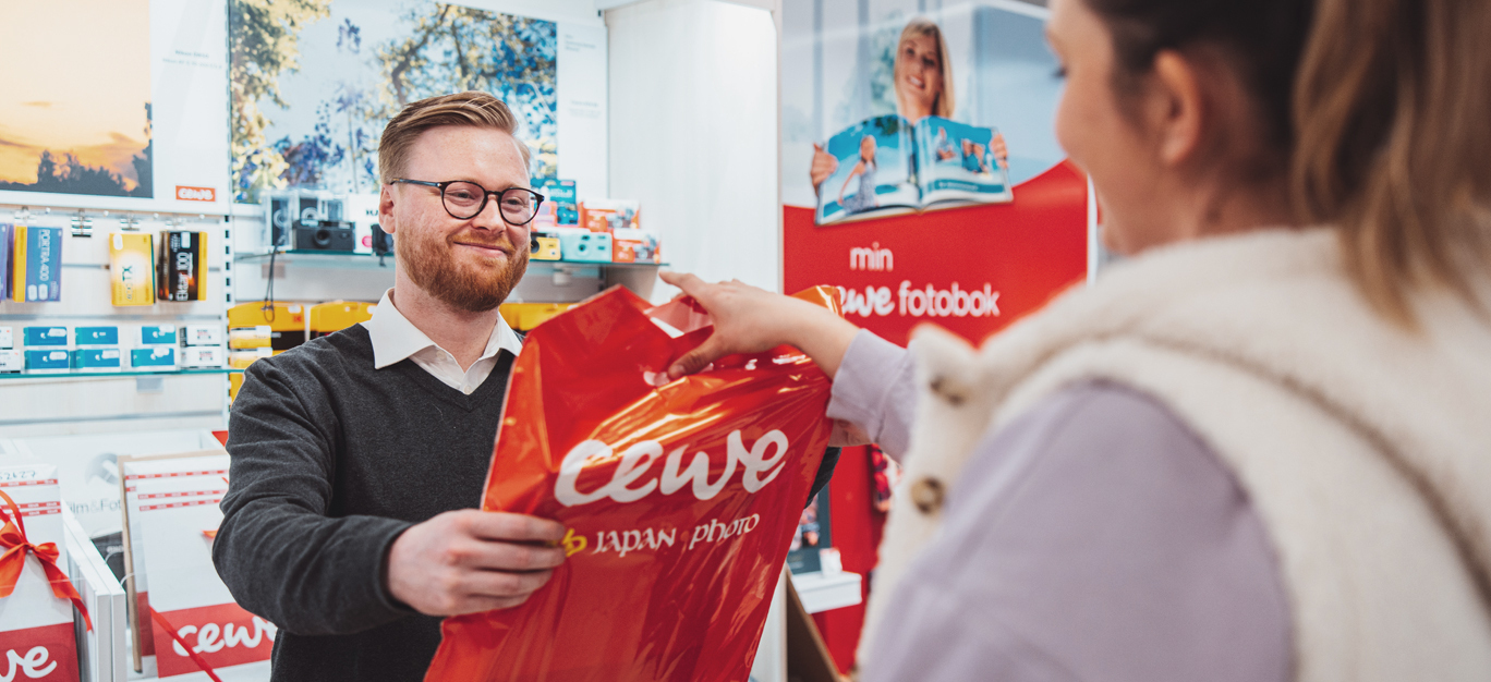 Kunde mottar en CEWE-pose fra en ansatt i en fargerik fotobutikk.