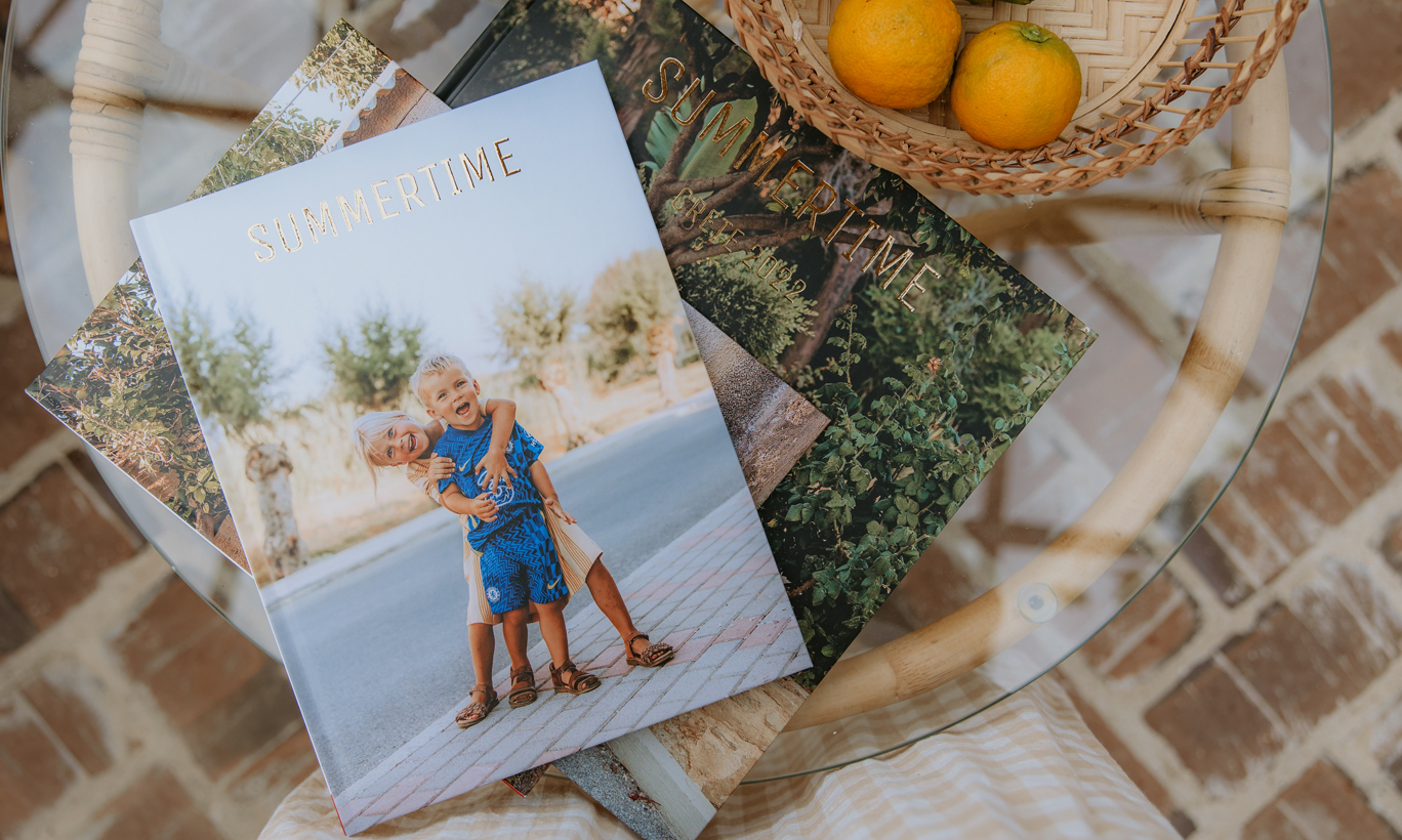 Fotobøker med teksten "Summertime" på et bord, ved siden av en kurv med ferske appelsiner.