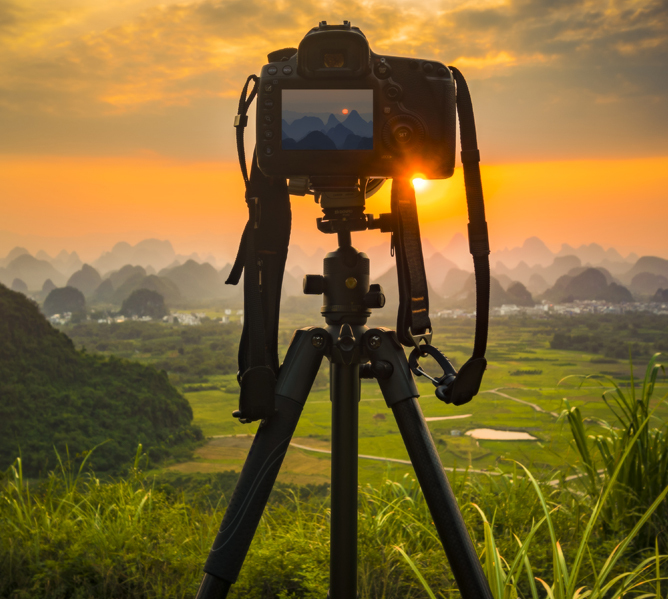 Et kamera på stativ som fanger en solnedgang over et grønt landskap med fjell i bakgrunnen Et kamera på stativ som fanger en solnedgang over et grønt landskap med fjell i bakgrunnen