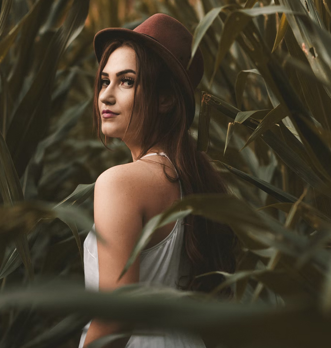 Kvinne med langt, brunt hår og hatt står i en åker omgitt av grønne planter.