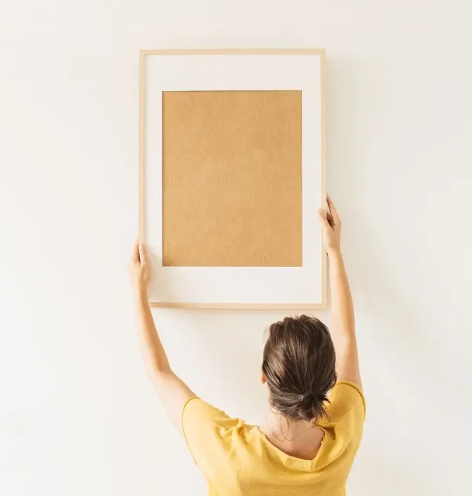 Kvinne med oppsatt hår henger opp en stor, tom bilderamme med trefarget kant på en hvit vegg