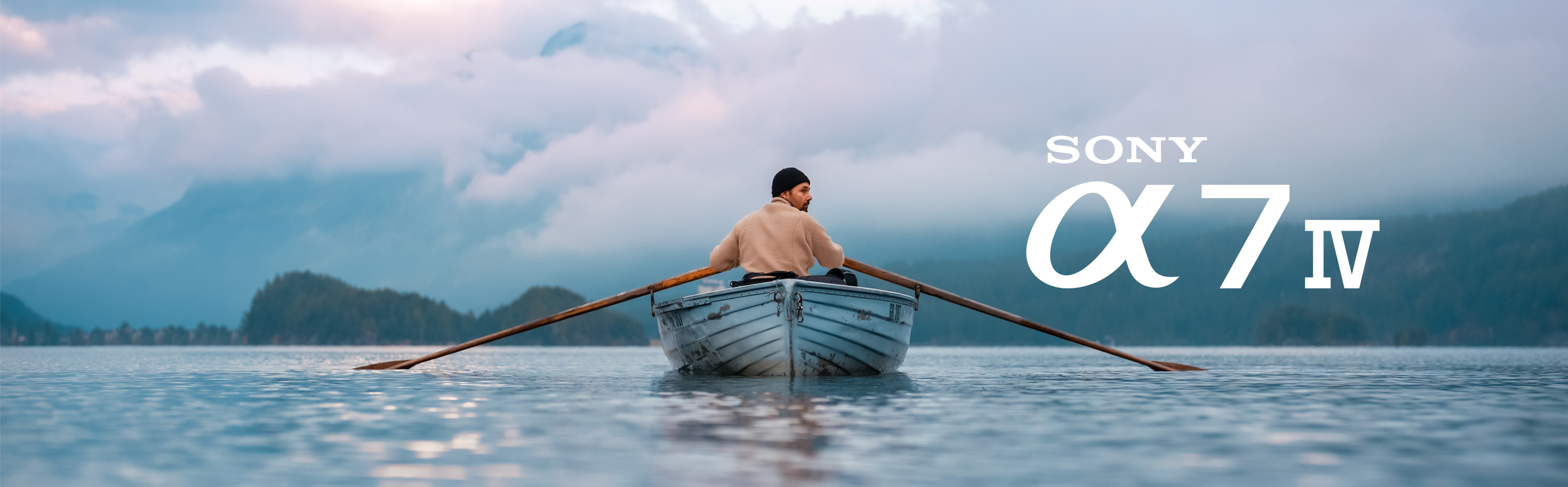 En person ror en båt på en stille innsjø omgitt av fjell og tåkedekte skyer