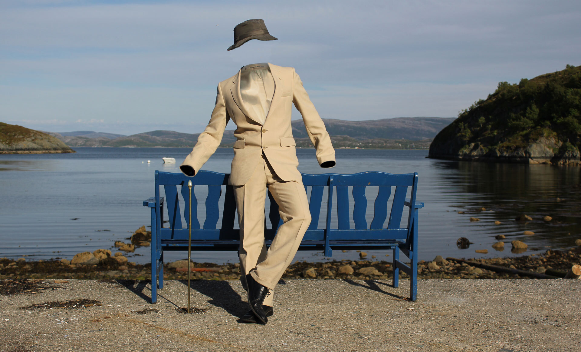 En beige dress uten person, med en hatt og stokk, lent mot en blå benk ved vannet i et naturskjønt landskap