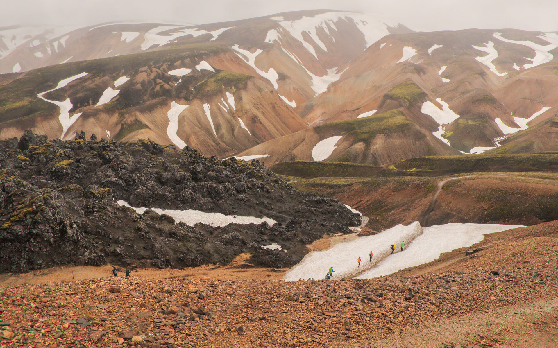 Fjellandskap med fargerike ryggformasjoner, lavaområder og snøflekker, vandrere krysser en liten snøsti i dalen
