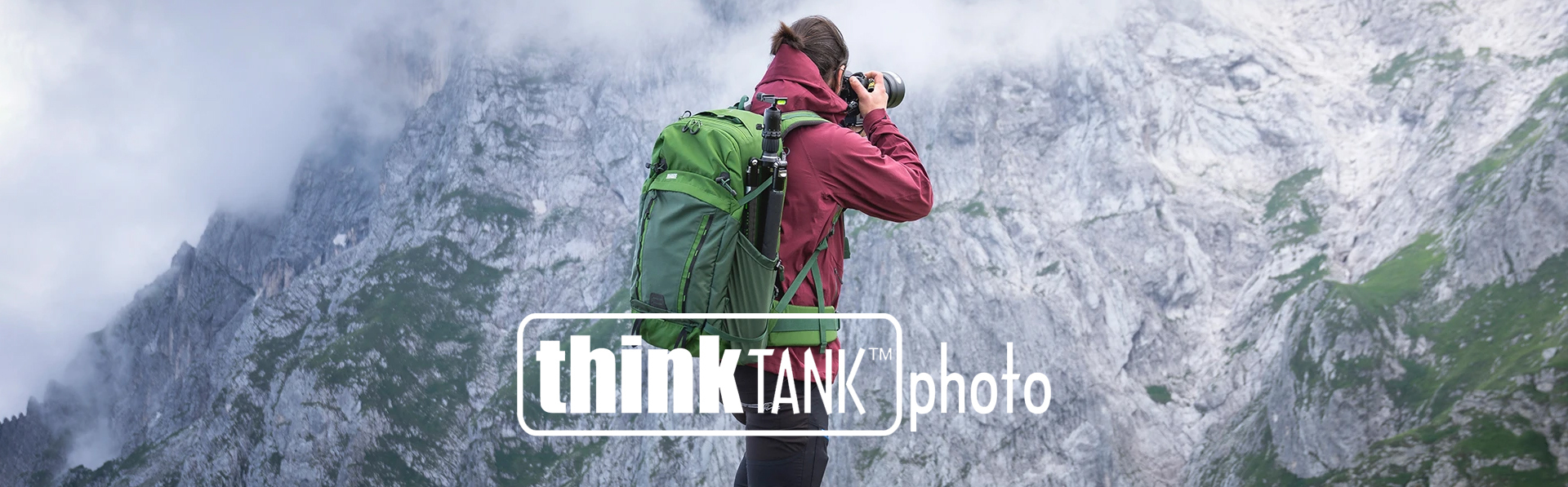 En person i rød jakke med en grønn ryggsekk fotograferer fjell innhyllet i tåke