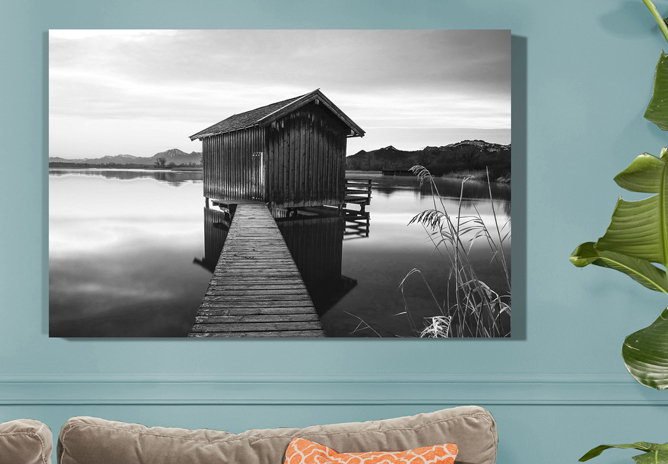 Et svart-hvitt bilde av en trebrygge som leder til en liten naustbygning på en stille innsjø