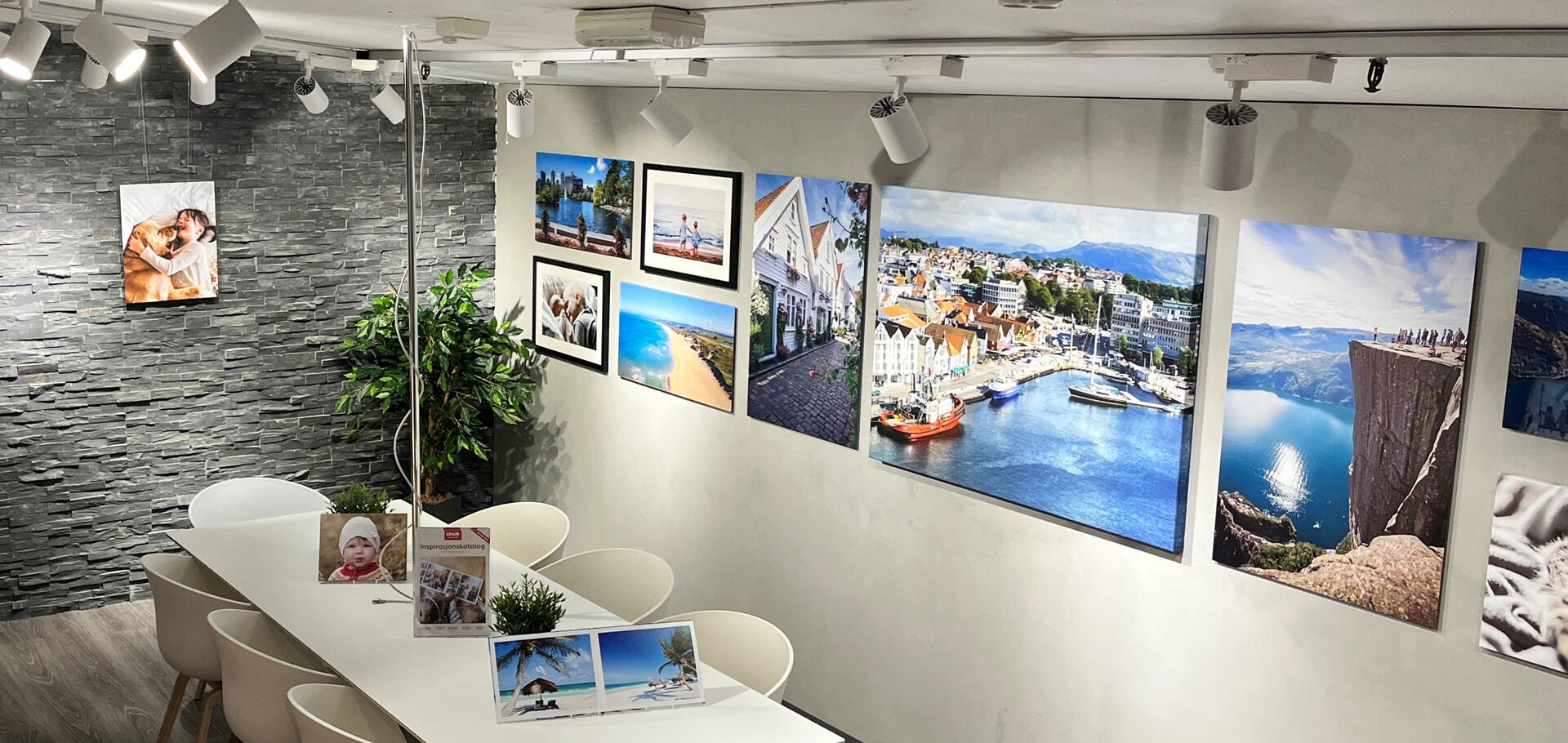 Lyse og moderne lokaler med grå steinvegg, en grønn plante og et hvitt bord med stoler; veggdekorasjon med fargerike fotografier av natur, byer og strender