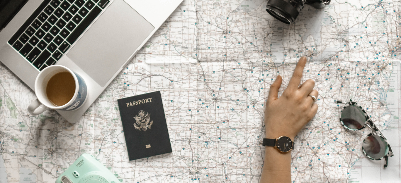 Hånd peker på et kart med en kopp kaffe, pass, kamera og solbriller rundt.