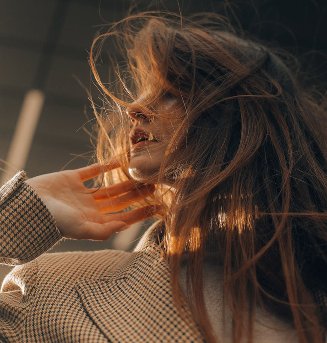 Person med langt, bølgete hår som blir opplyst av varmt sollys, iført en rutete jakke. Hånden løftet mot håret.