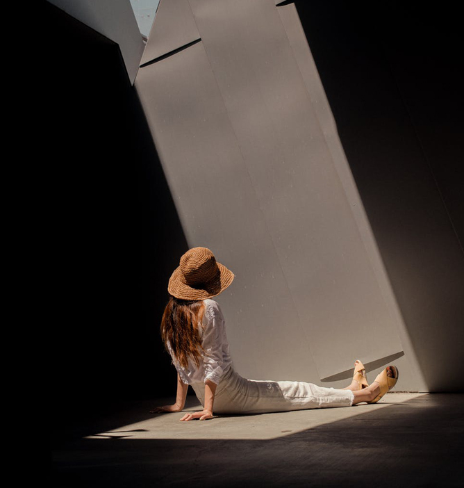 Kvinne med stor stråhatt og hvite klær sitter på gulvet i sollys som strømmer gjennom en geometrisk åpning Kvinne med stor stråhatt og hvite klær sitter på gulvet i sollys som strømmer gjennom en geometrisk åpning