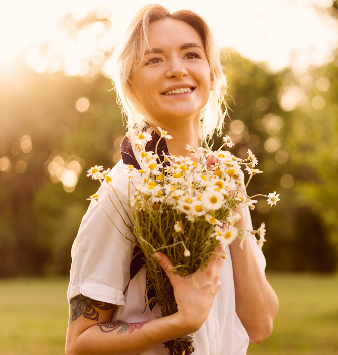 Kvinne i solnedgang holder en bukett med prestekrager i begge hender, omgitt av grønt gress og trær. Kvinne i solnedgang holder en bukett med prestekrager i begge hender, omgitt av grønt gress og trær.