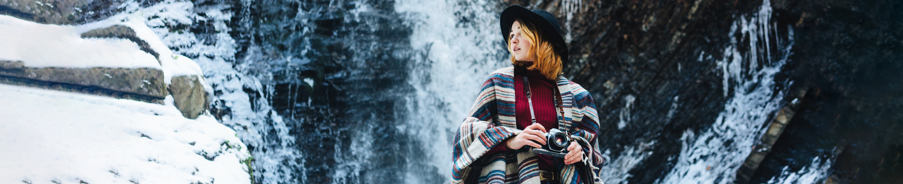 Person med stripete poncho og svart hatt står foran en frossen foss og holder et kamera.