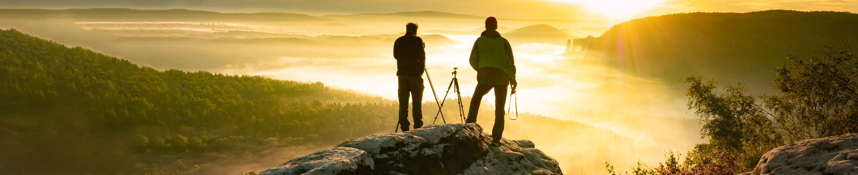To personer på en fjelltopp med kamera stativ ser på soloppgangen over en tåkelagt dal med grønne åser og gyllent lys To personer på en fjelltopp med kamera stativ ser på soloppgangen over en tåkelagt dal med grønne åser og gyllent lys