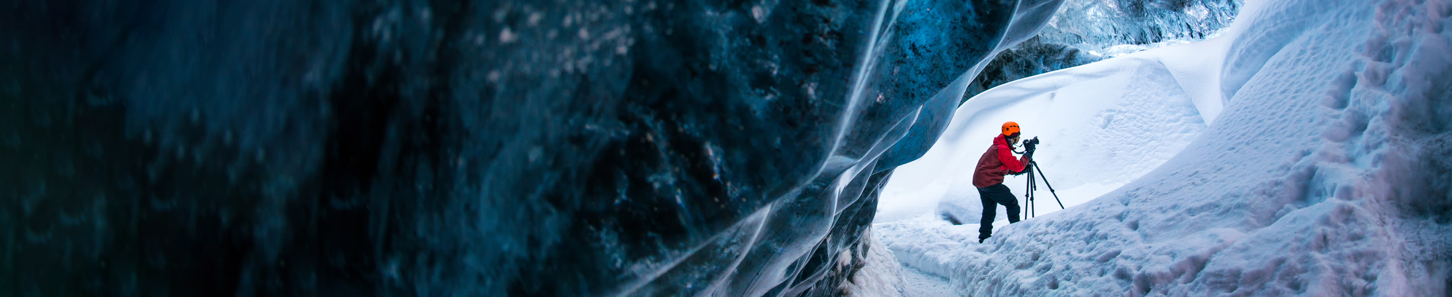 Person i rød jakke og oransje hjelm tar bilder med stativ i snødekt isgrotte