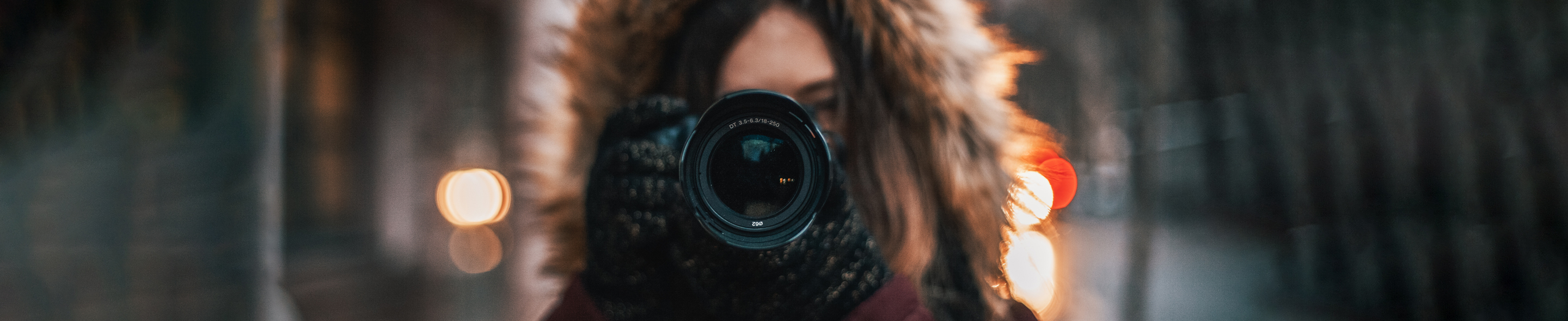 Person med vinterjakke og pelshette står ute om kvelden med uskarpe bylys i bakgrunnen