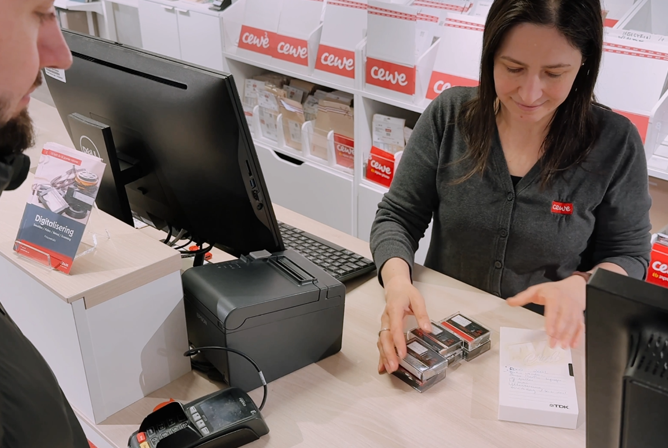 En kunde ved kassen i en CEWE-butikk, der en ansatt hjelper med å klargjøre fotokassetter ved disken En kunde ved kassen i en CEWE-butikk, der en ansatt hjelper med å klargjøre fotokassetter ved disken