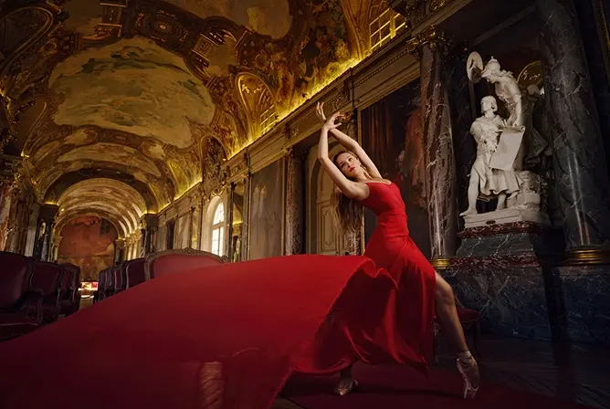 En person i en rød kjole som danser grasiøst i en overdådig sal dekorert med marmorsøyler, statuer og detaljerte takmalerier