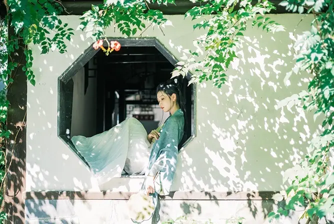 Person i tradisjonell japansk drakt står ved et oktagonalt vindu omgitt av grønne blader.